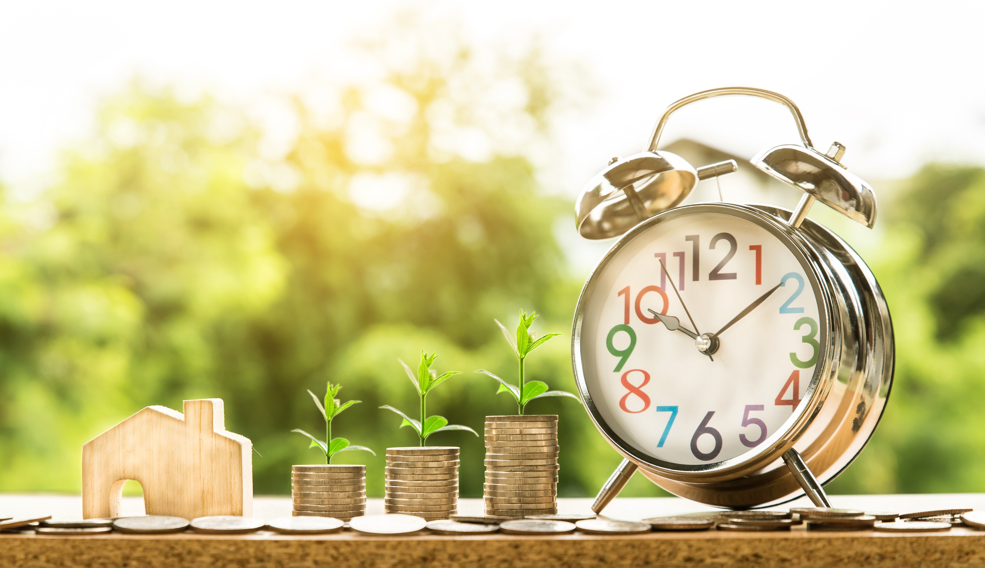 This is a photo of an alarm clock with three small piles of gold coins with new plants growing out of them and a small wooden house.